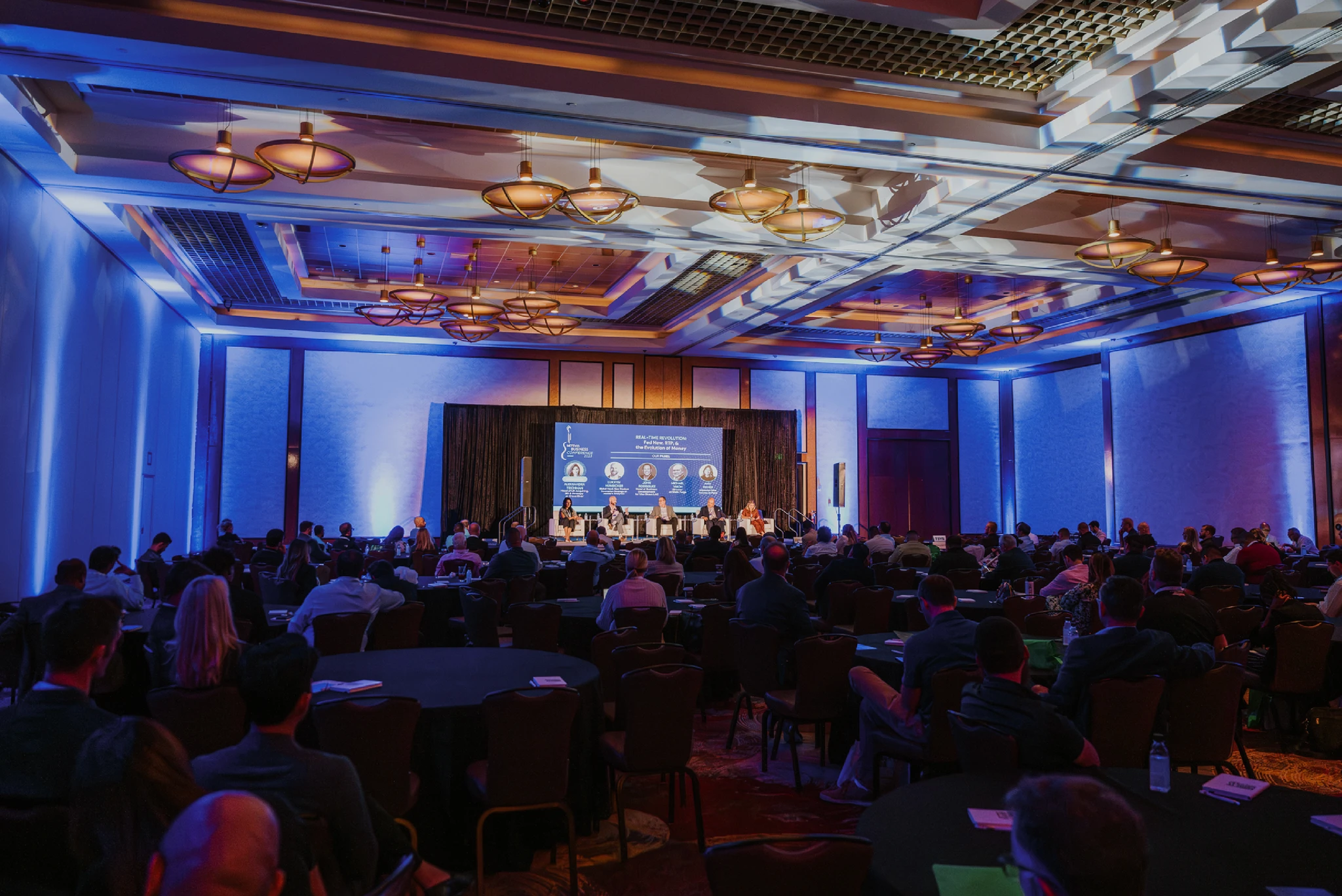 Conference audience facing a panel in a blue-lit ballroom